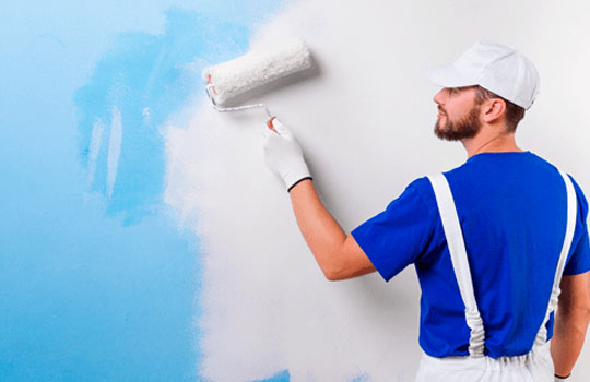 A person in a blue shirt and white suspenders House Painter Service a wall with a roller.
